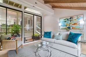 Ein modernes Wohnzimmer mit einem weißen Sofa, einem Glastisch und durch große Glasschübe Blick auf einen Außenbereich mit Pflanzen.