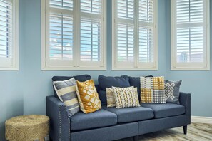 Ein blaues Sofa mit gemusterten Kissen vor einer hellblauen Wand, das von Fenstern mit weißen Rahmen gesäumt wird.