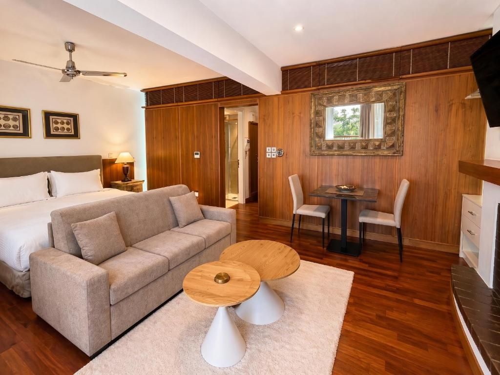 A hotel room with a bed, a sofa, a dining table with chairs, a ceiling fan, and a large mirror on the wall.