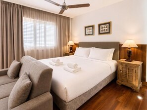 A hotel room with a bed, two bedside lamps, a ceiling fan, a sofa, and a wooden cabinet. - Ithaki Beach Apartment by TrulyCyprus (Lemesos)