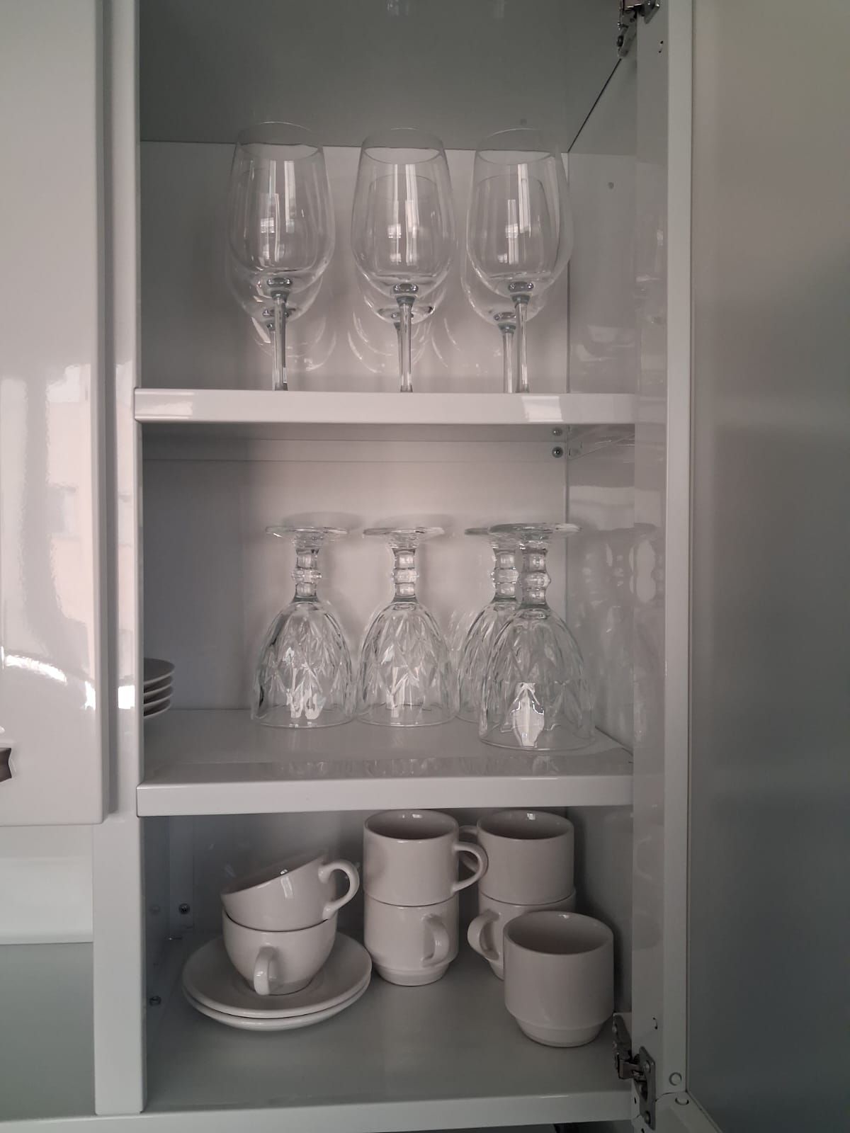 Un mueble blanco con estantes que contienen copas de vino, botelleros de cristal y tazas blancas.