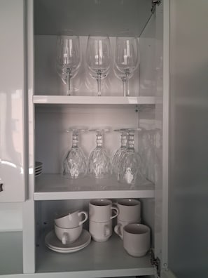 Un mueble blanco con estantes que contienen copas de vino, botelleros de cristal y tazas blancas.