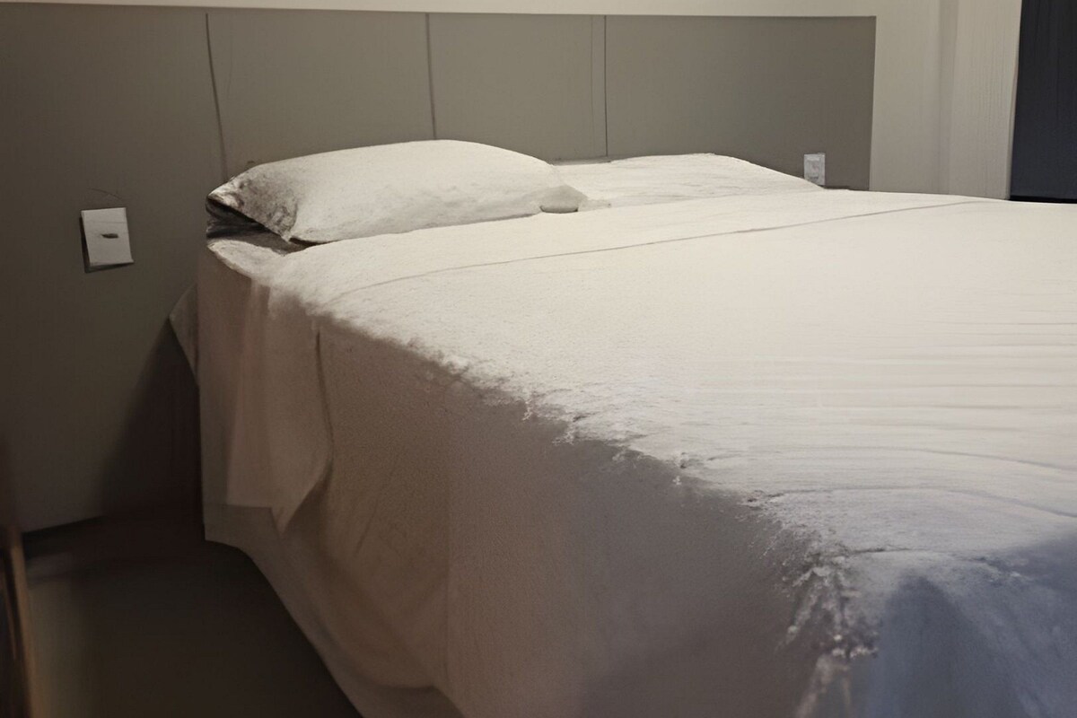 Una cama individual con colchón y almohadas blancas, un cabecero e interruptor de luz en la pared.