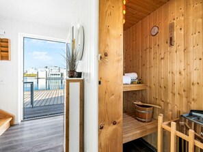 Une cabine de sauna en bois, avec un banc en bois, une petite étagère où sont posées des serviettes et des bouteilles, et une horloge au mur.