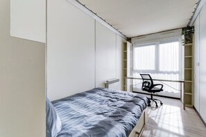A bedroom with a bed, a desk, and a chair. There is a window with blinds and a radiator. - 77 - Le Lokup - Wifi - Proche tram (Reims)