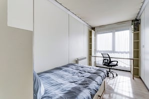 Ein Schlafzimmer mit Bett, Schreibtisch und Stuhl. Ein Fenster mit Jalousien und eine Heizung sind ebenfalls vorhanden.