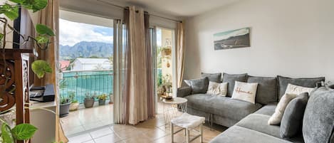 A living room with a grey sofa, a small table, and a balcony with potted plants.