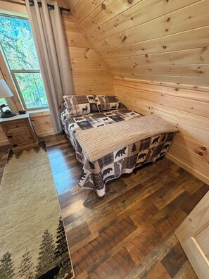 A wooden cabin bedroom with a bed, a nightstand, a lamp, and a view of the forest. - New 3-bedroom cabin in ADK with hot tub (Wells)