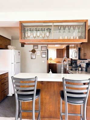 A kitchen with a central island, two bar-style chairs, a refrigerator, and a glass-fronted cabinet with decorative items. - Hillside Haven Cozy 2BR Near Pullman and WSU (Colfax)