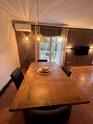 Et spiseområde med et træbord, stole, en hængende lampe og et fjernsyn monteret på væggen.