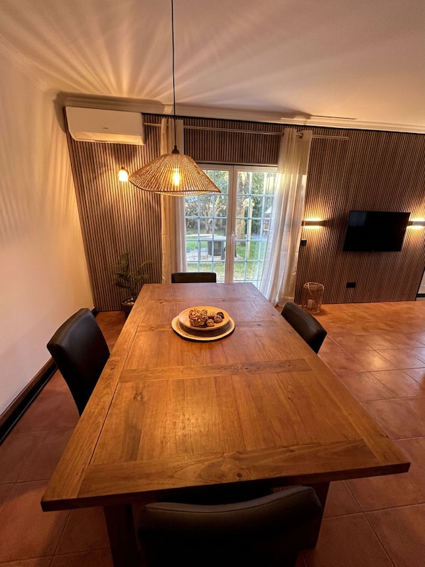 Ein Essbereich mit einem Holztisch, Stühlen, einer hängenden Leuchte und einem an der Wand montierten Fernseher.