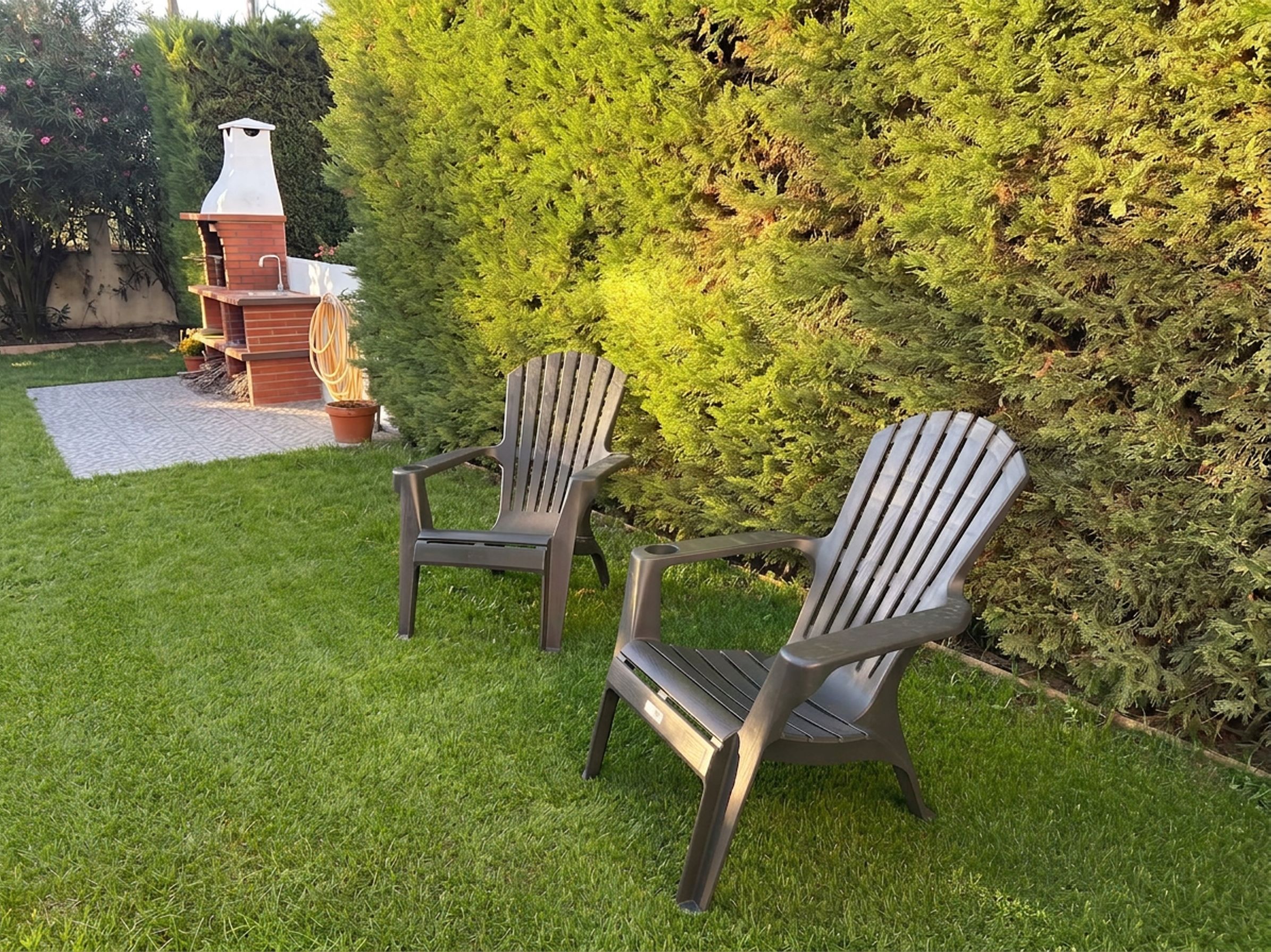 Deux fauteuils Adirondack dans une cour arrière verdoyante, avec un barbecue en briques et une haie.