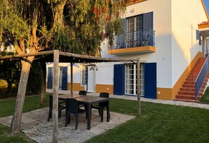 Un espacio al aire libre techado con mesa y sillas, una pérgola de madera y un edificio con contraventanas azules y un balcón.