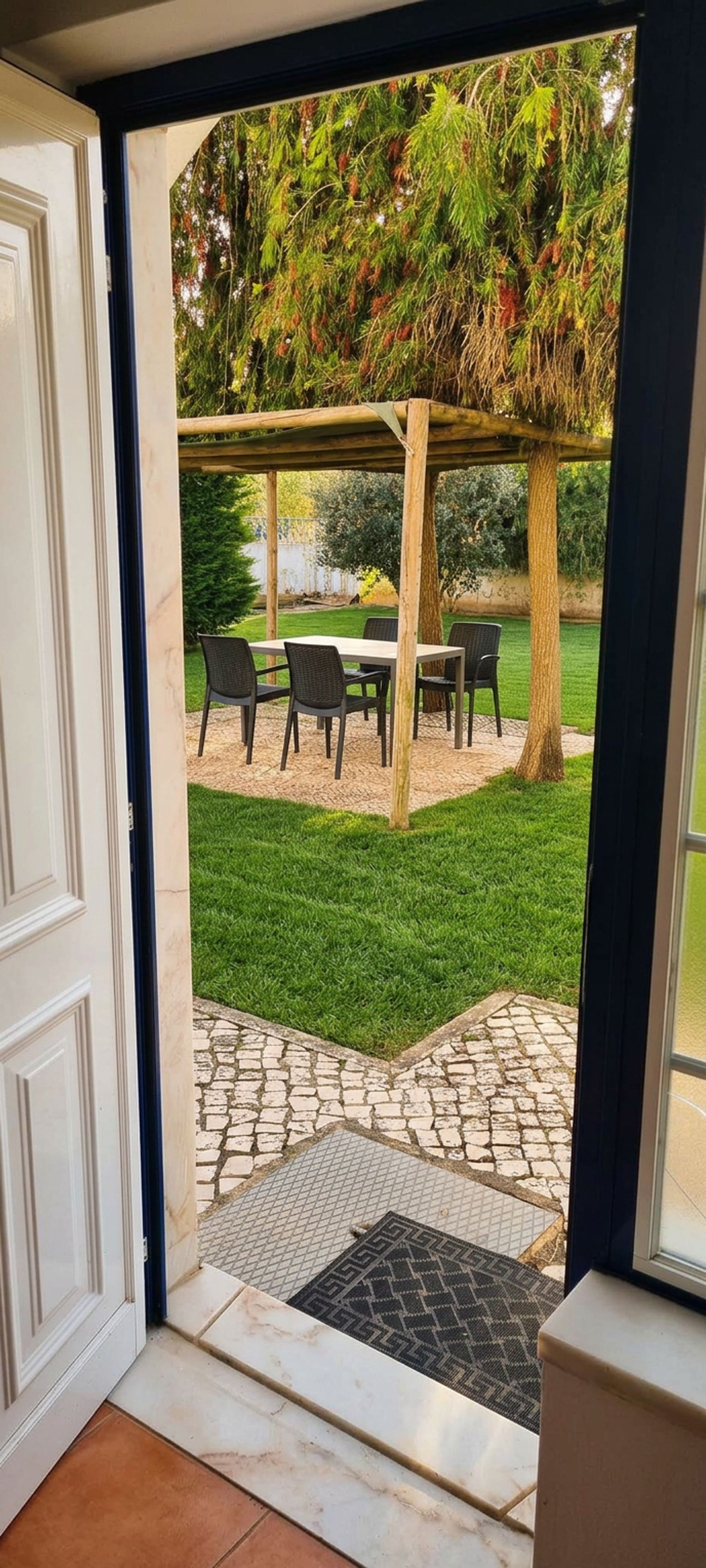 En vy genom en dörröppning in till en uteplats med en pergola, utomhusmöbler och en gång av sten.