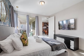 A modern hotel room with a bed, a TV, a desk, and a view of a cityscape. - 11br Near Disney Pool, spa & Home Theater (Kissimmee)