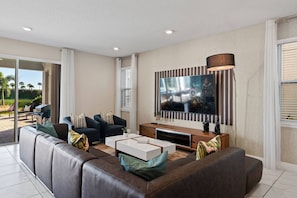 A modern living room with a large flat-screen TV, comfortable seating, and a view of the outdoors. - 11br Near Disney Pool, spa & Home Theater (Kissimmee)