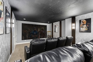 A home theater room with black leather reclining seats, a large flat-screen TV, and framed movie posters on the walls. - 11br Near Disney Pool, spa & Home Theater (Kissimmee)