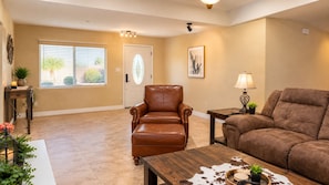 A spacious living room with a brown leather armchair, a brown sofa, and a wooden coffee table. There is a door with a glass panel, a window with blinds, and a framed picture on the wall. - Relax, Recharge & Play at 3-bedroom Bluewater Bungalow (Lake Havasu City)