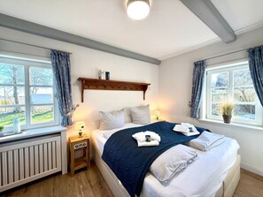 Ein Hotelzimmer mit Bett, Nachttischen, Heizkörper, einem Fenster mit Vorhängen und einem Regal über dem Bett.