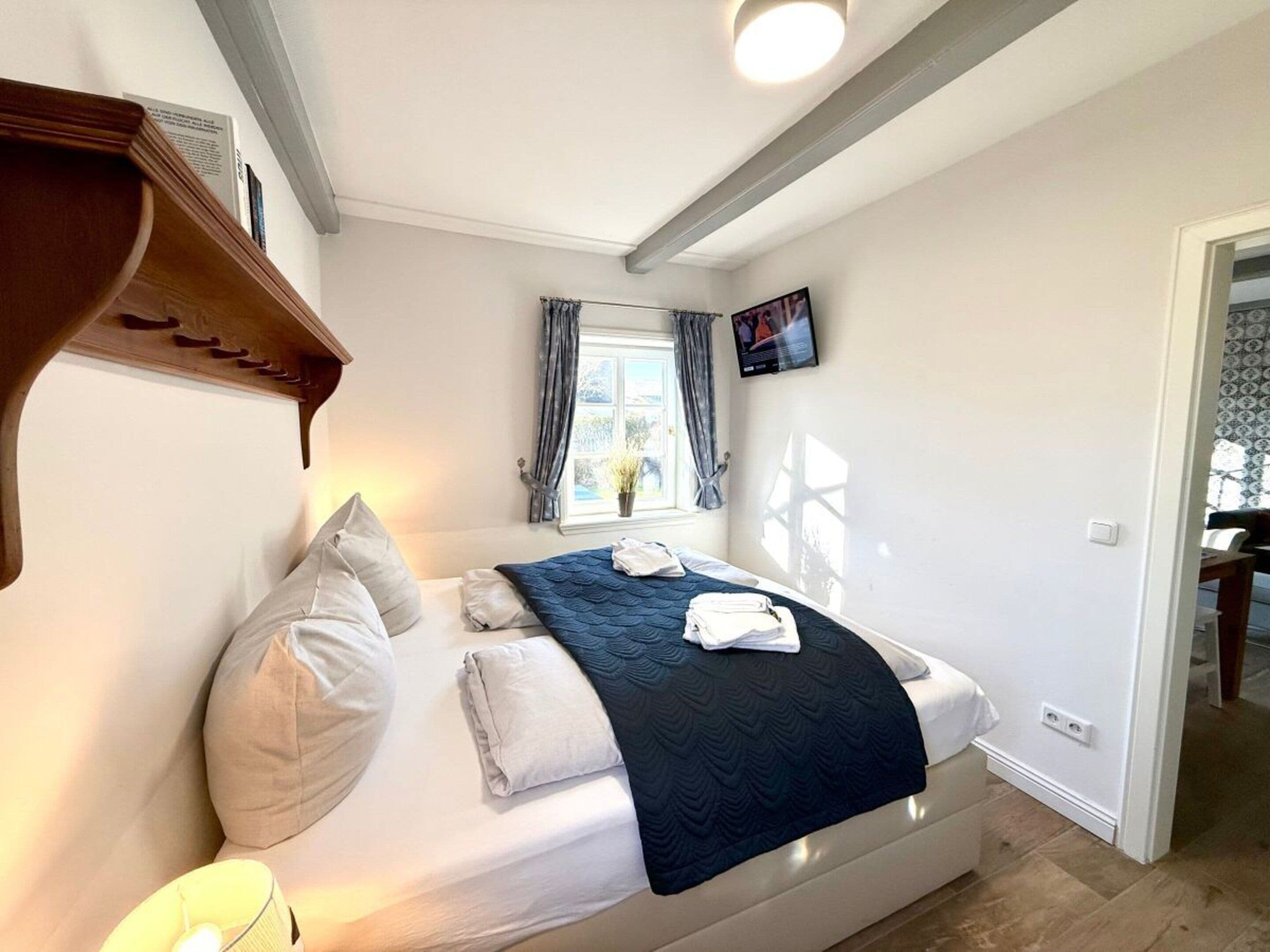 Un dormitorio con cama, almohadas, una colcha, televisor y ventana con cortinas.
