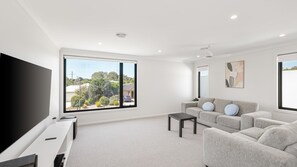 A modern living room with a large TV, grey sofa, and a view of a residential area. - The Gathering House – Spacious Island Retreat (Cowes)