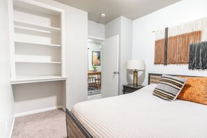 Een slaapkamer met een bed, nachtkastje, lamp en een wanddecoratie van macramé.