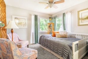 Quarto com cama, duas cadeiras, janela com cortinas, ventilador de teto e um quadro na parede.
