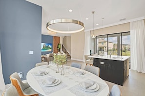 A modern dining area with a round table set for six, a kitchen island, and a view of the outside. - 5BR Near Disney Private Pool & Lake View (Davenport)