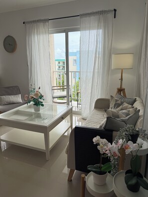 A modern living room with a glass coffee table, a sofa, a bed with pillows, and a view of a building through sheer curtains. - 2-bedroom apartment in marvelous Punta Cana with cool AC (Punta Cana)