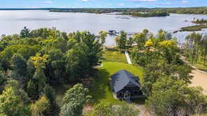 A house with a dark roof, surrounded by a well-maintained lawn, near a large body of water. - Pelican Lake Getaway, 2BR + Office, Beach & Pedal Boat (Breezy Point)