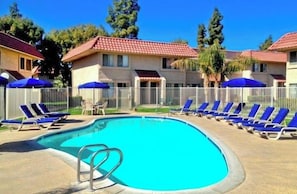 A swimming pool with lounge chairs and umbrellas, surrounded by a fence and buildings with red-tiled roofs. - Walk-to-Head-Trip Indio Retreat (Indio)