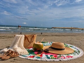 Eine Strandansicht mit Sonnenhut, Sonnenbrille, einem Kokosdrink und einer geflochtenen Tasche auf einer bunten Decke.