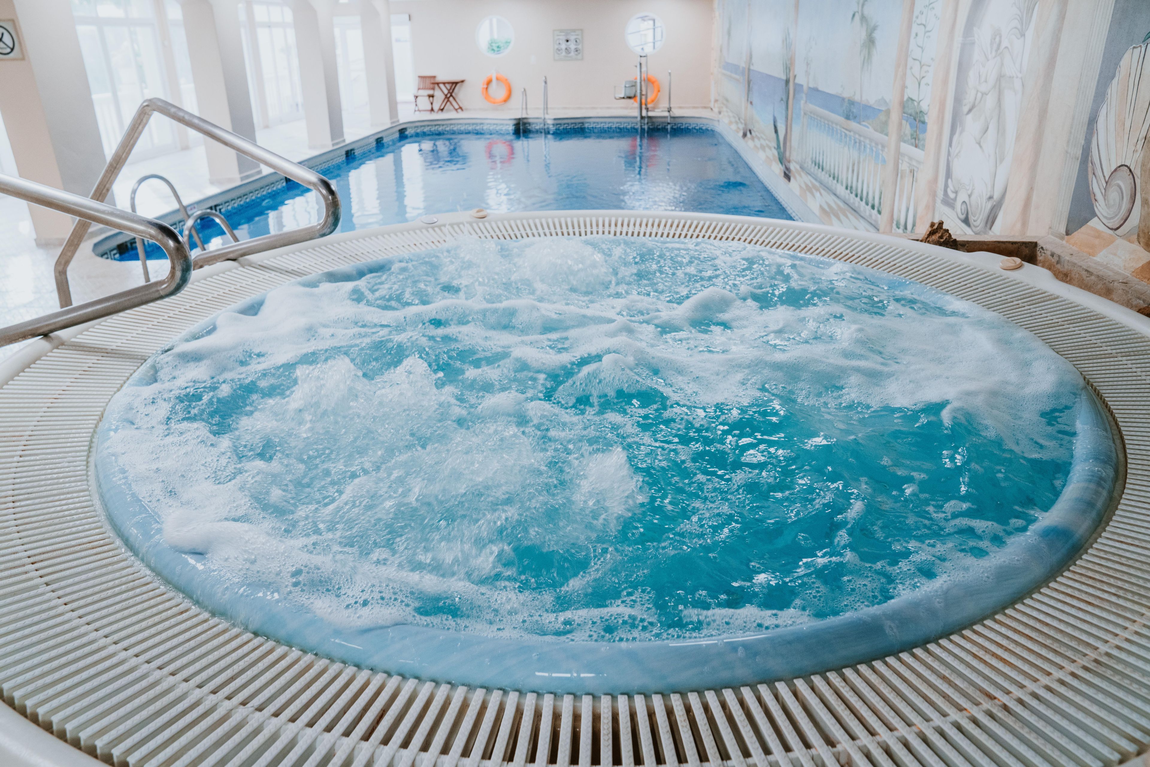 Un jacuzzi con una escalera metálica, rodeado de un suelo con baldosas y una piscina al fondo.