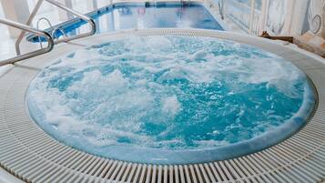A hot tub with a metal ladder, surrounded by a tiled floor, and a pool in the background.