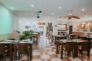 A restaurant with a checkered floor, wooden tables, and chairs. There are potted plants and a television screen on the wall. - Crown Resorts (Mijas)