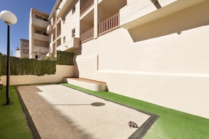 Un patio con césped artificial verde, una zona de juegos de hormigón y un edificio con balcones.