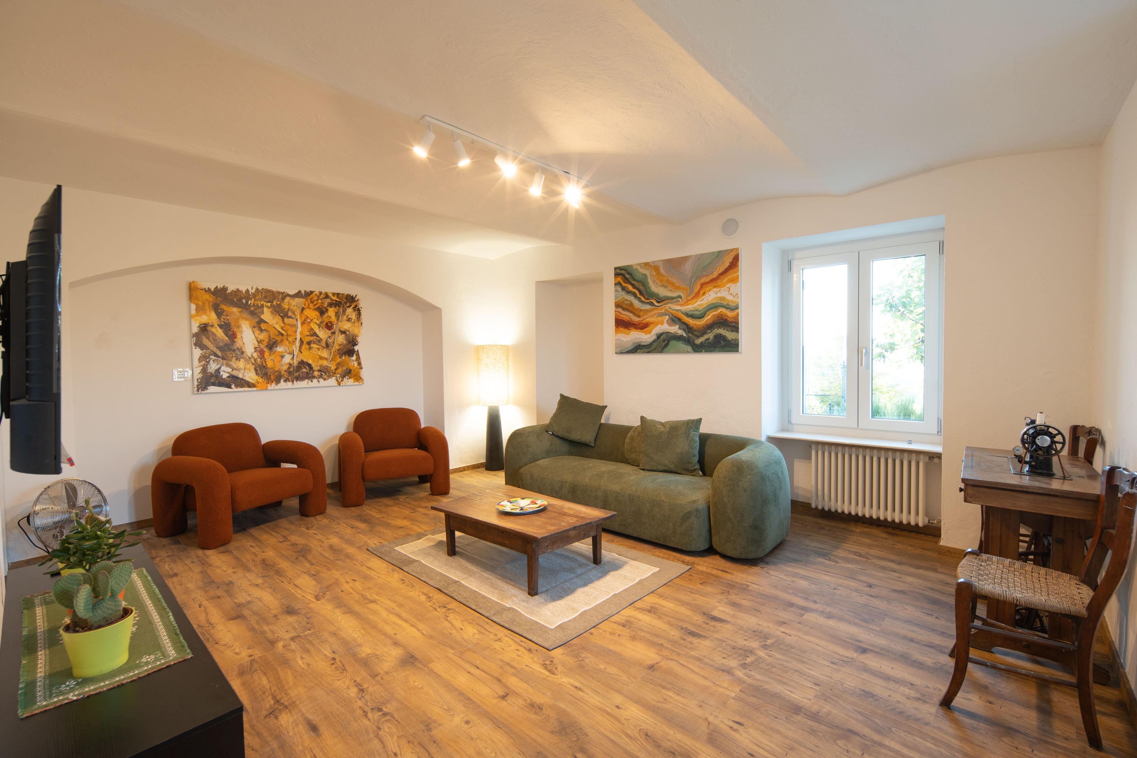 A living room with a sofa, two armchairs, a coffee table, and a lamp. There are abstract paintings on the walls and a window with a view.