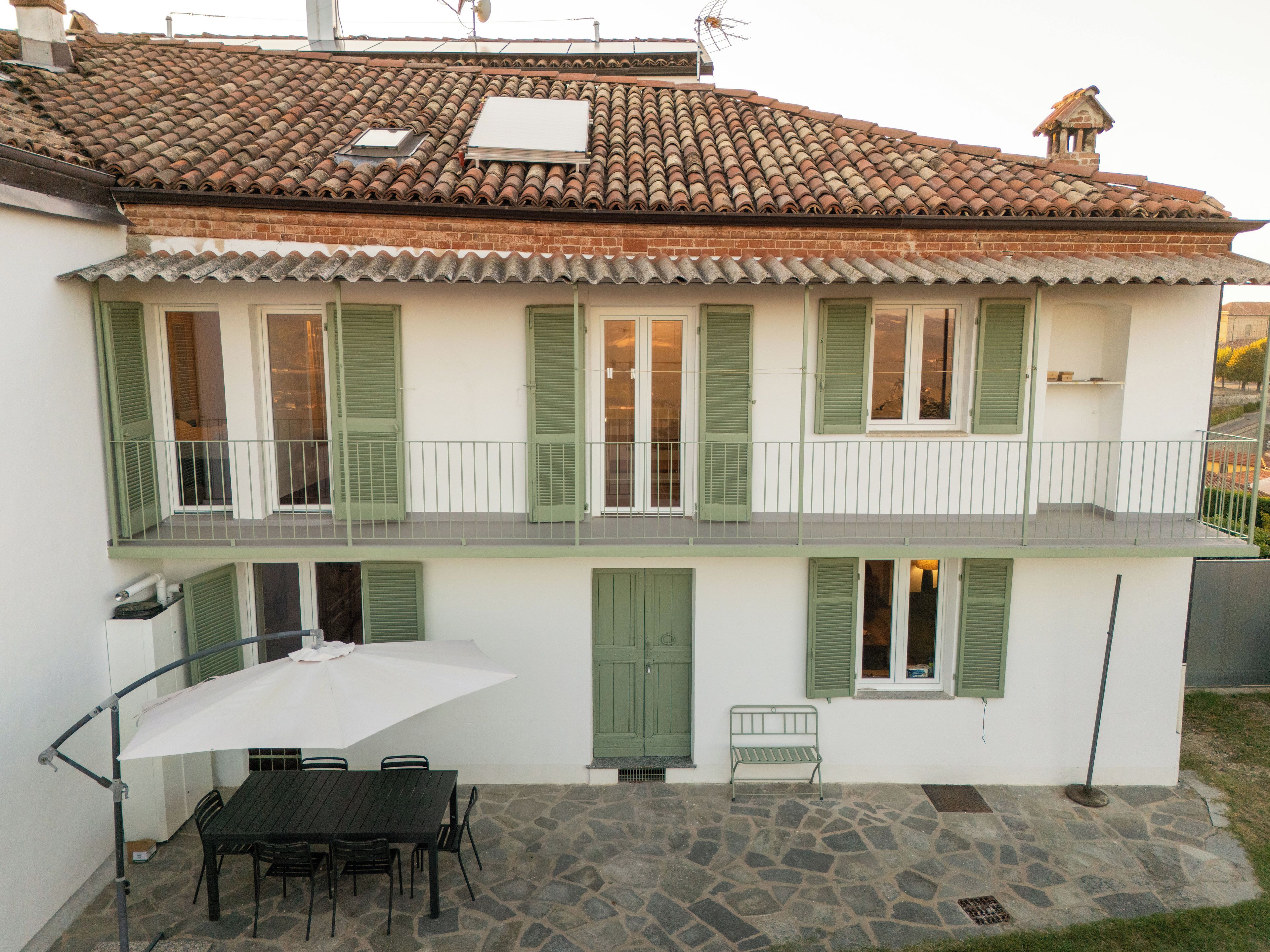 Un edificio a due piani con tetto in tegole, persiane verdi e un patio in pietra con tavolo e sedie.