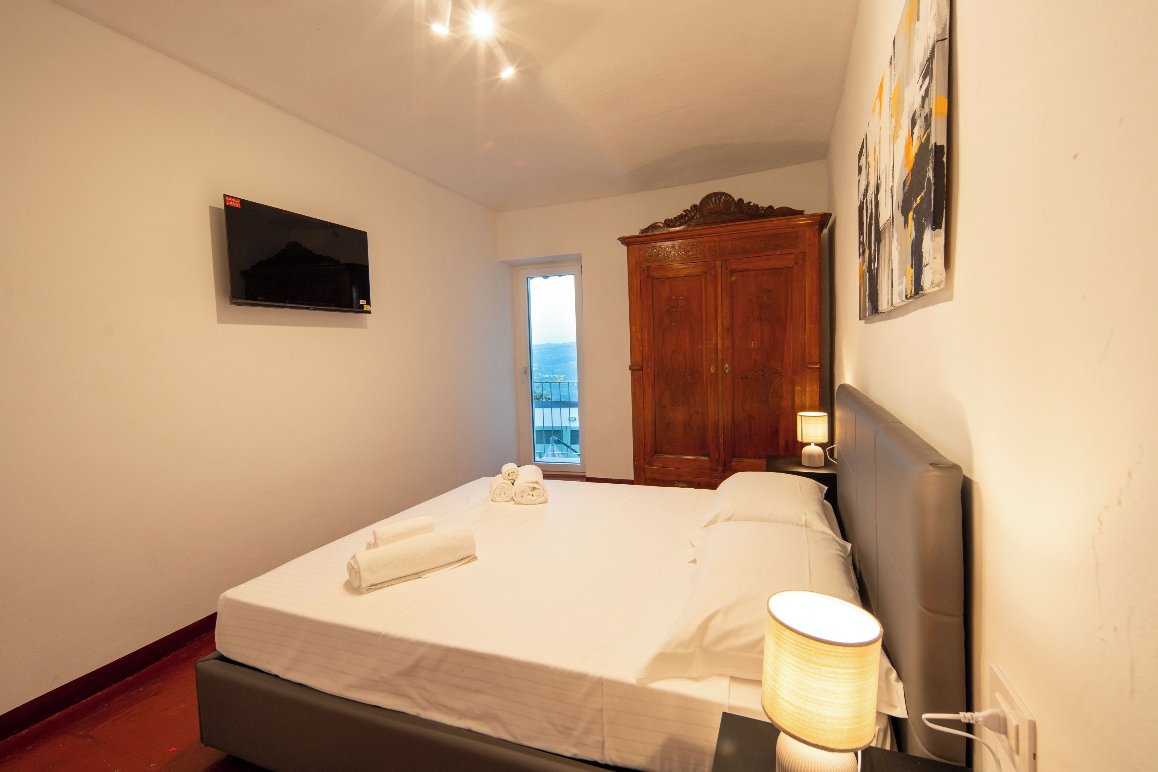 A hotel room with a bed, a TV mounted on the wall, a wooden wardrobe, and a window with a view.