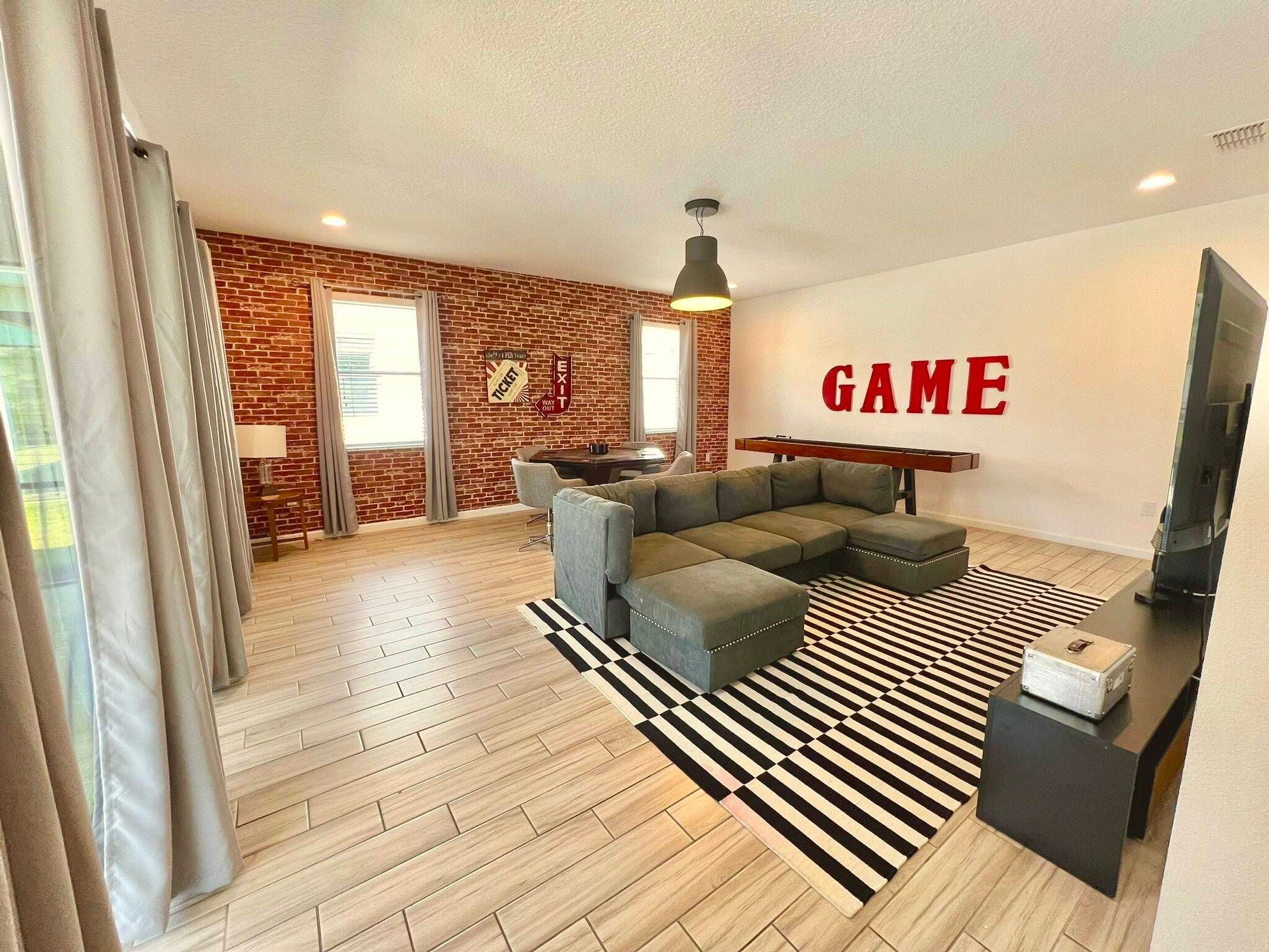 Un salon moderne avec un canapé d’angle, un tapis rayé noir et blanc, un téléviseur et un mur sur lequel est inscrit le mot « GAME » en lettres rouges.