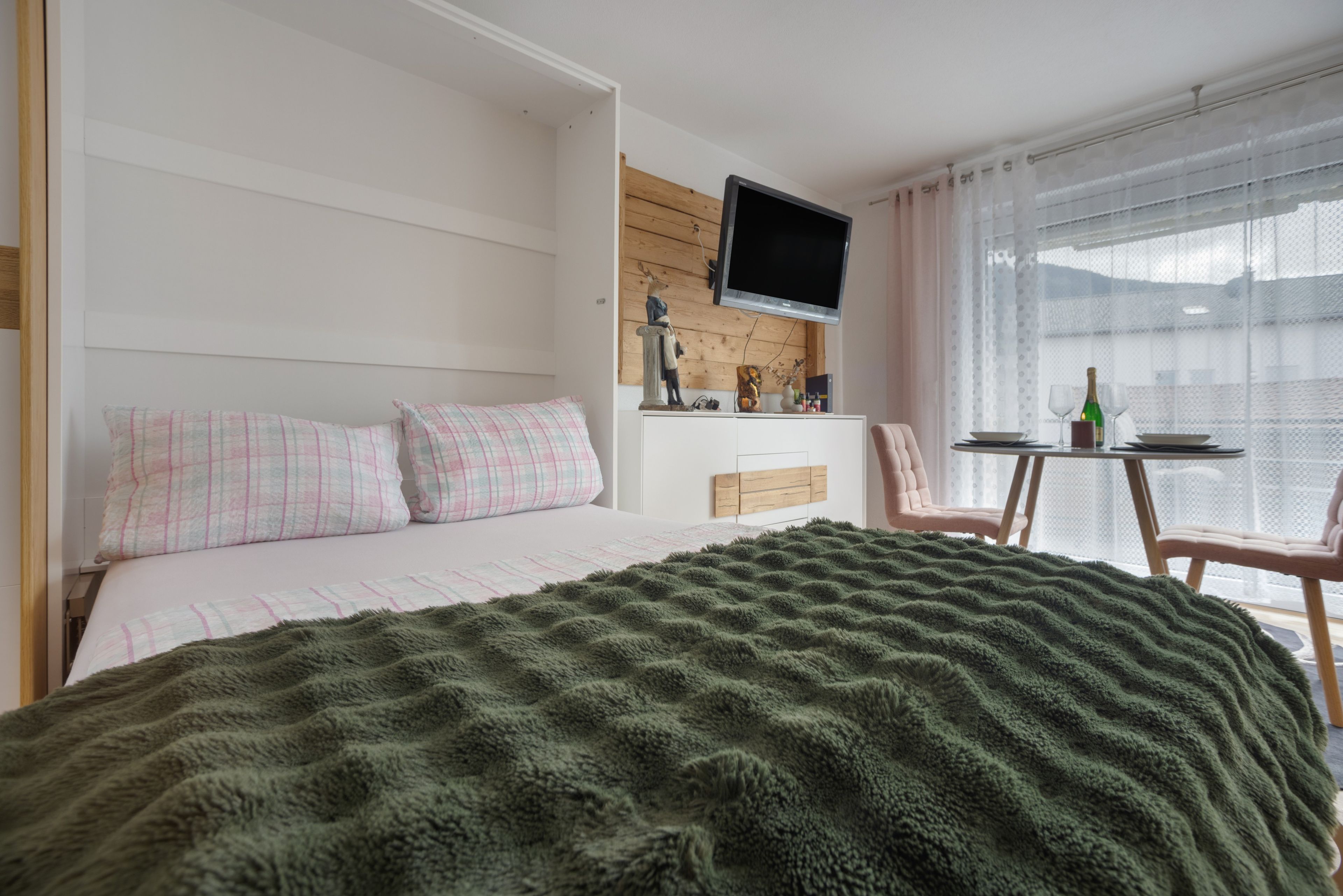 Une chambre à coucher avec un lit, un téléviseur fixé au mur, une petite table avec des chaises et une fenêtre avec des rideaux.