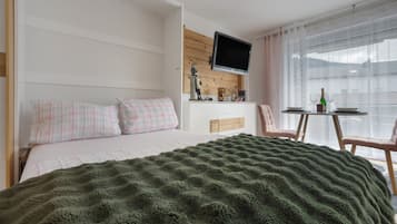 Une chambre à coucher avec un lit, un téléviseur fixé au mur, une petite table avec des chaises et une fenêtre avec des rideaux.