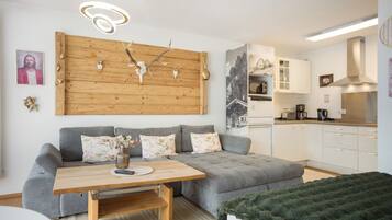 Un salon moderne avec un canapé gris, une table basse en bois et un coin cuisine doté de meubles blancs et d’une cuisinière.