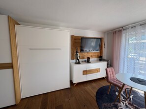 Et moderne værelse med et hvidt skab, et træskab med et fjernsyn, et lille bord med en stol og et vindue med gardiner.