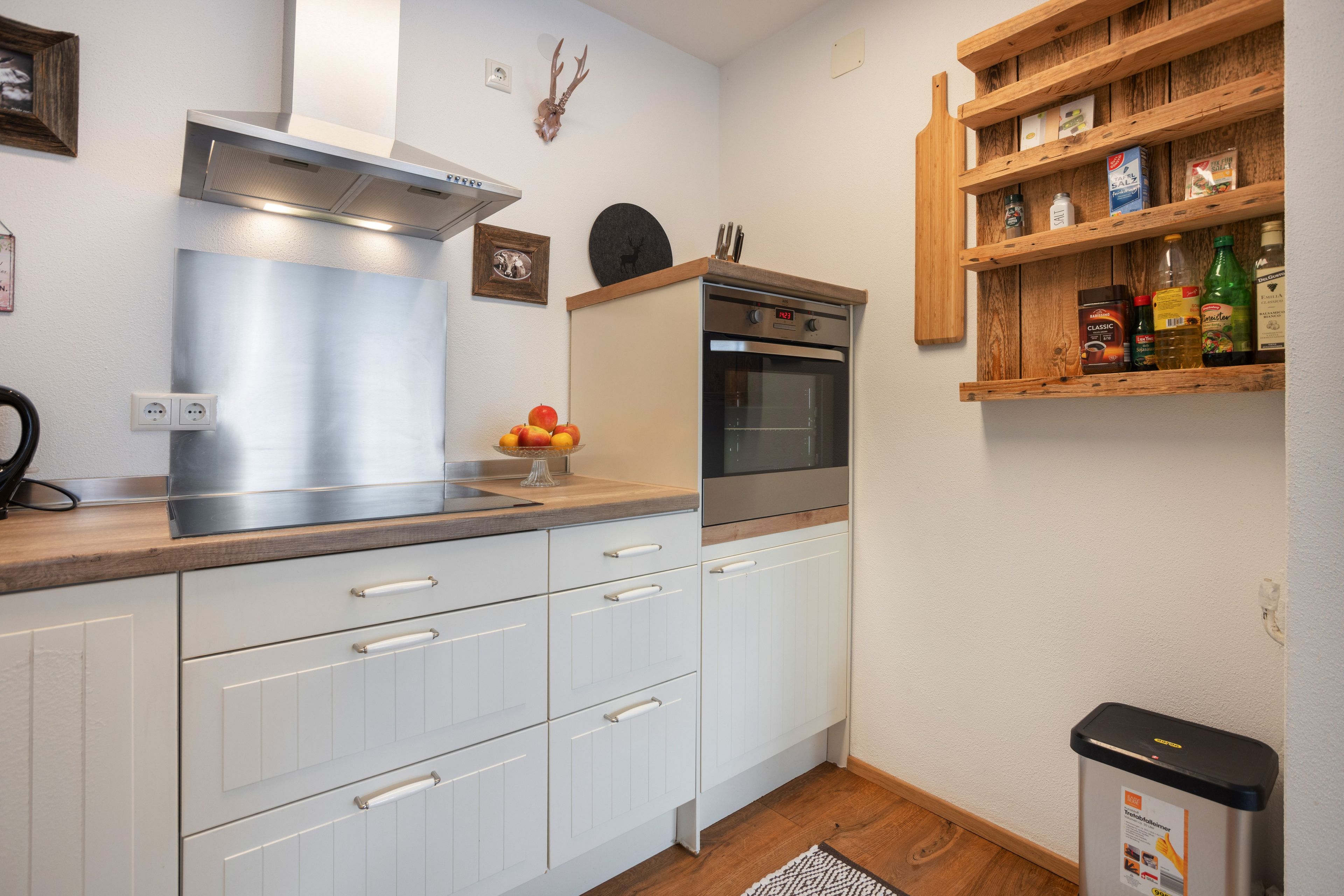 A compact kitchen with white cabinets, a stainless steel range hood, a built-in oven, and a wooden shelf with various items.