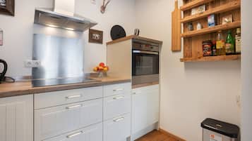 A compact kitchen with white cabinets, a stainless steel range hood, a built-in oven, and a wooden shelf with various items.