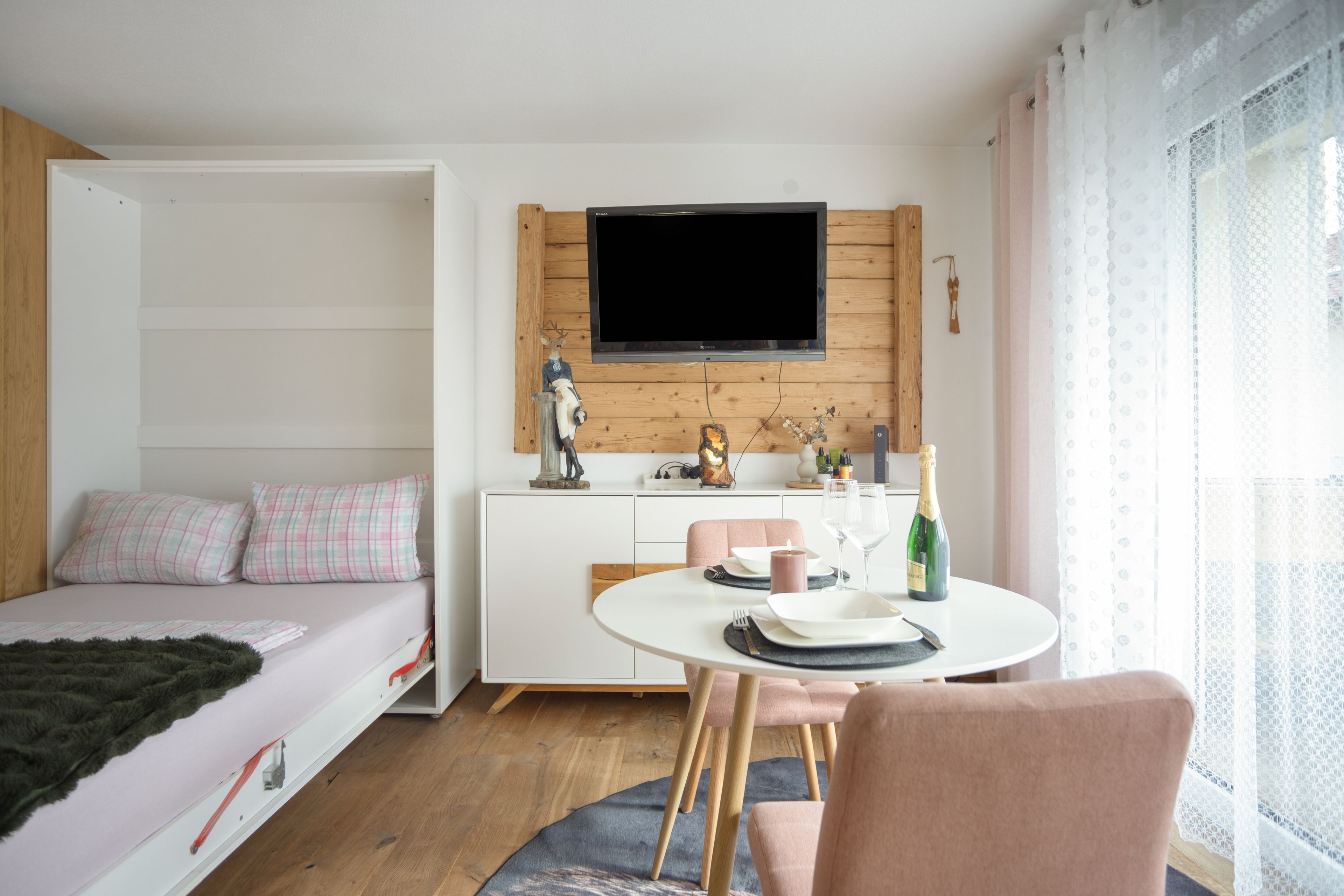 Une chambre moderne comprenant un lit, un coin repas avec une table et des chaises, un téléviseur à écran plat fixé à un mur en bois, et une fenêtre agrémentée de rideaux légers.