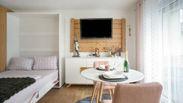 Une chambre moderne comprenant un lit, un coin repas avec une table et des chaises, un téléviseur à écran plat fixé à un mur en bois, et une fenêtre agrémentée de rideaux légers.