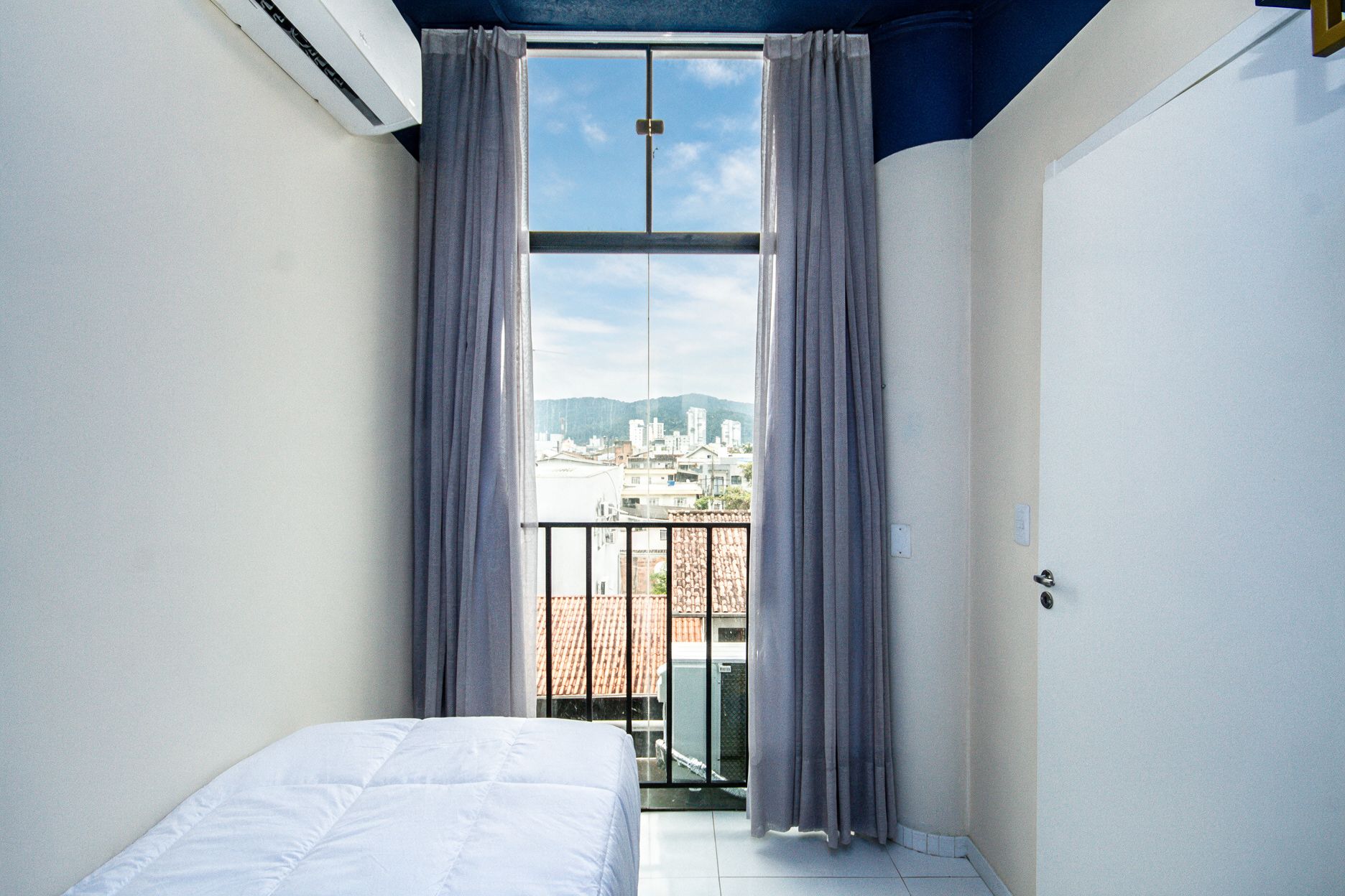 A room with a bed, a window with curtains, and a view of a cityscape.