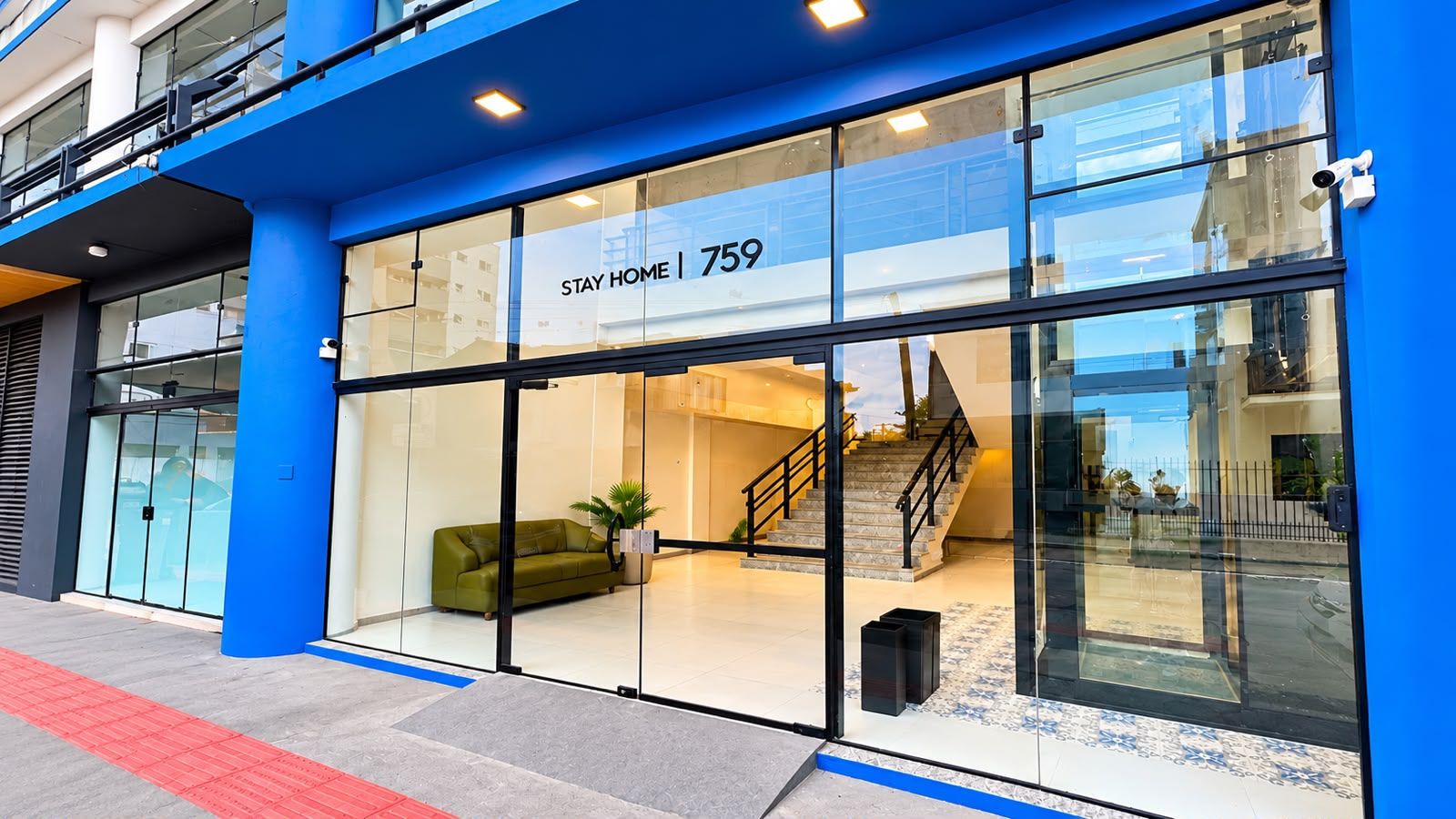 A modern hotel entrance with a glass facade, a green sofa inside, and a staircase leading to the upper floor.